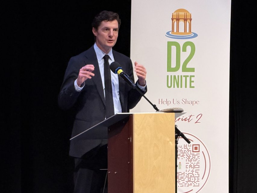 A man in a suit speaks at a podium with a microphone, standing next to a "D2 Unite" banner promoting District 2.