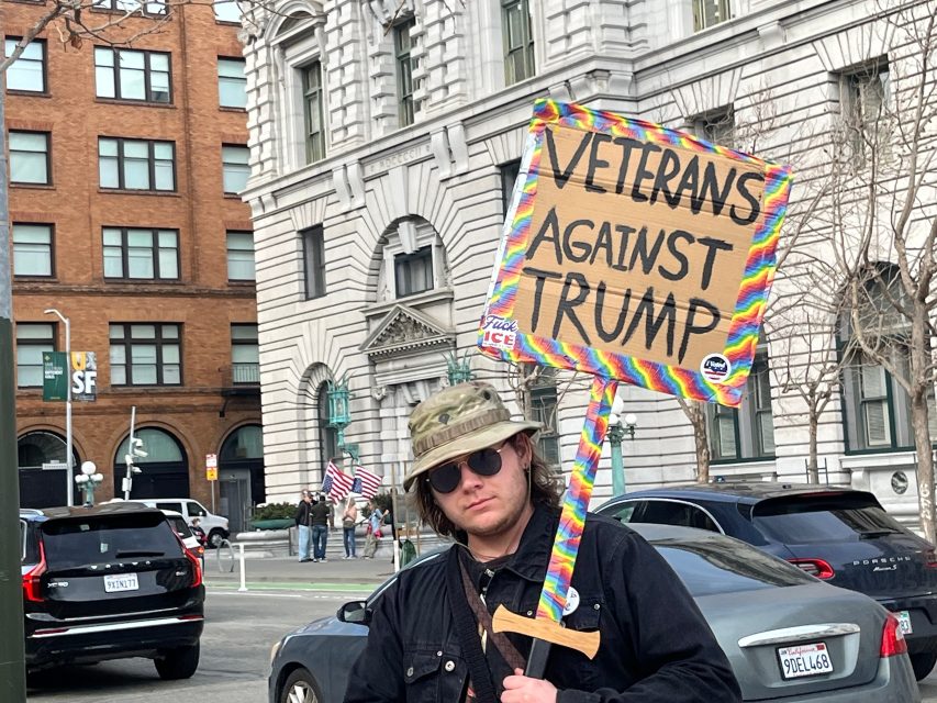 A person wearing sunglasses and a camouflage hat holds a colorful sign that reads "Veterans Against Trump" on a city street with buildings and cars in the background.