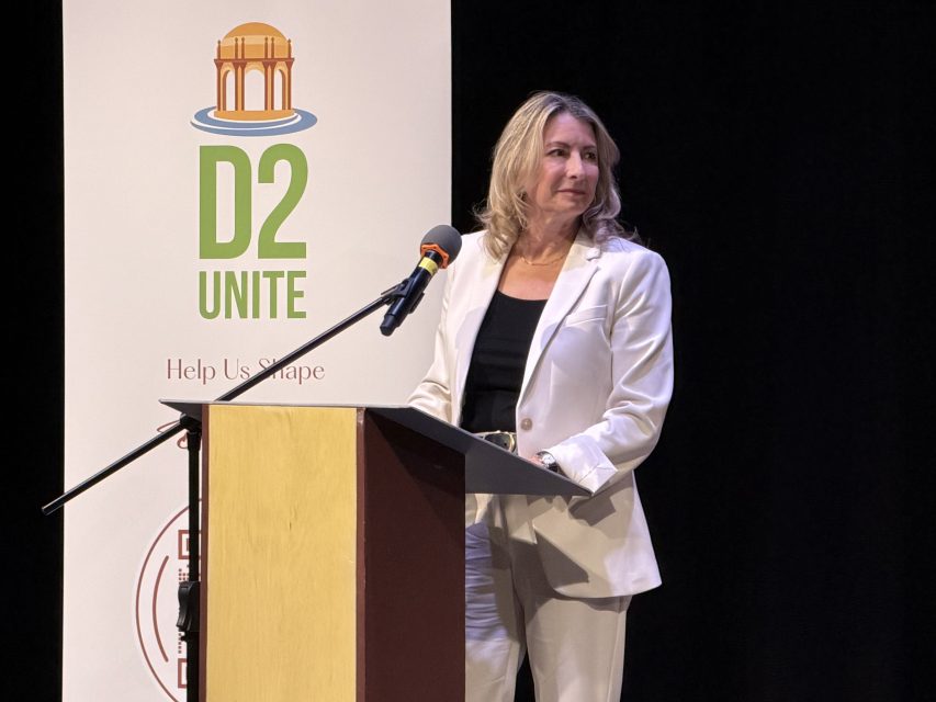 A woman in a white suit stands at a podium with a microphone, speaking at an event with a banner labeled "D2 UNITE" in the background.