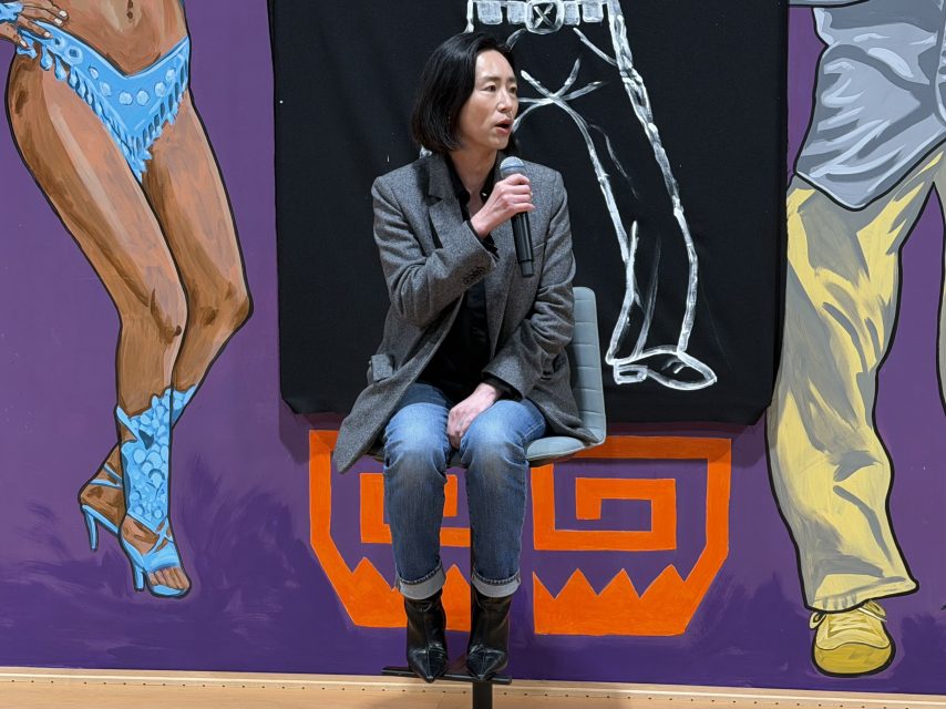 A person in a gray blazer and jeans sits on a stool, speaking into a microphone, in front of a colorful mural featuring painted legs and decorative patterns.