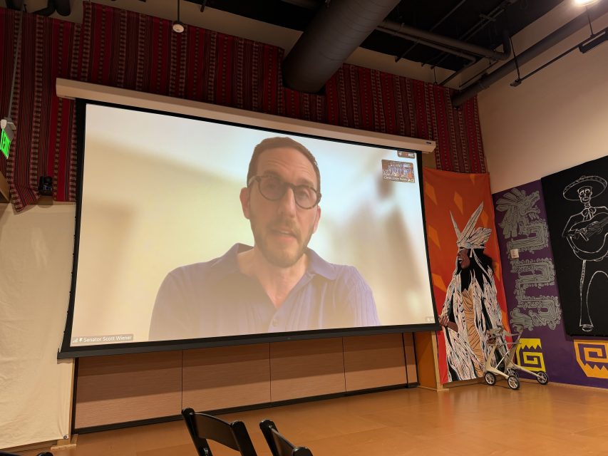 A large screen in a room displays a man speaking via video call. The room has patterned walls, artwork, chairs, and visible ceiling ducts.
