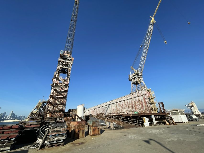 Two large industrial cranes stand on a concrete dock with rusted metal structures and a clear blue sky in the background.