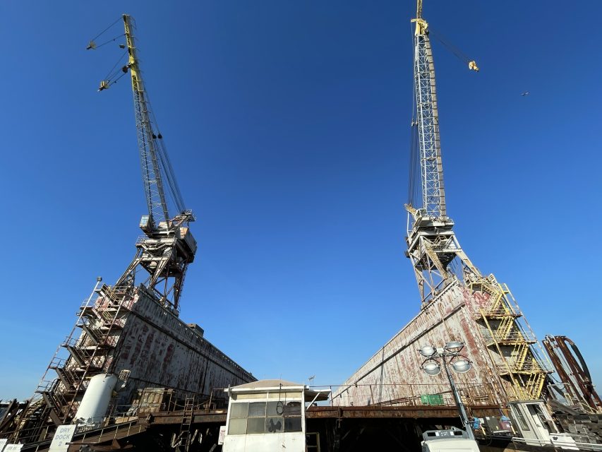 Two large, rusted industrial cranes with angled supports rise against a clear blue sky at a shipyard or dock facility, with equipment and buildings in the foreground.