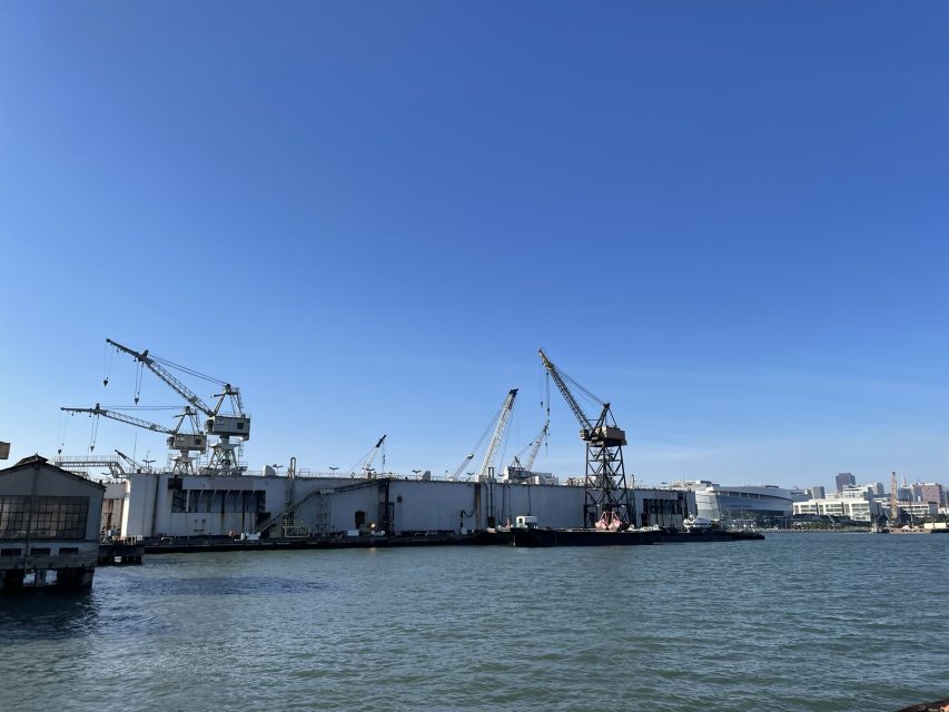 Industrial cranes and large buildings line the waterfront under a clear blue sky, with calm water in the foreground.