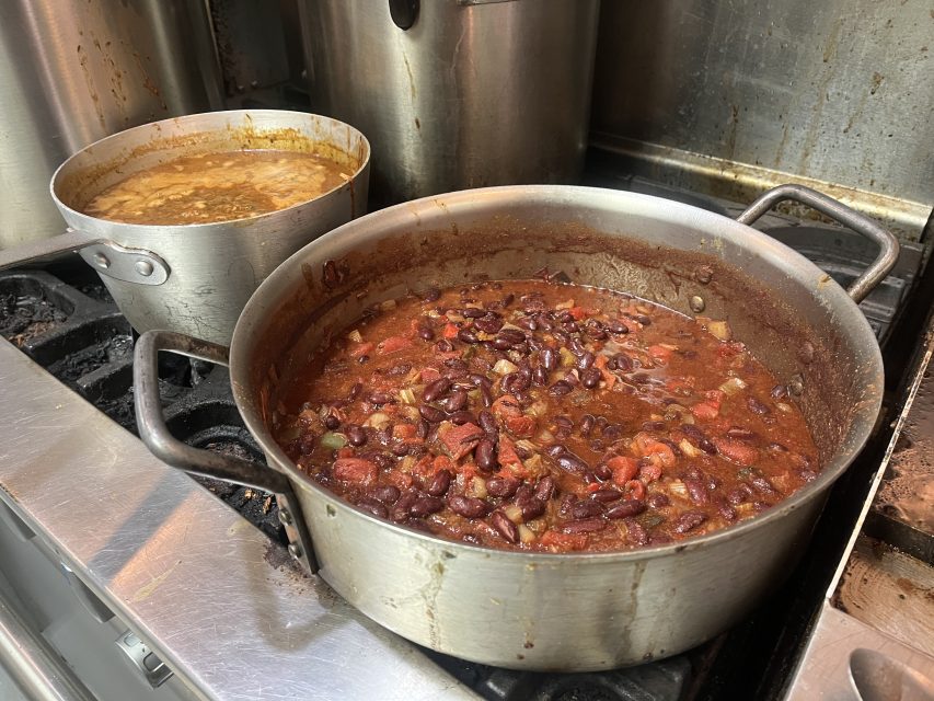 Two pots on a stovetop: one with red bean chili, the other with a lighter-colored soup or stew, both simmering in a commercial kitchen setting.