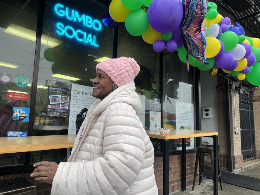 A woman in a pink hat and white coat smiles outside Gumbo Social restaurant, decorated with colorful balloons and Mardi Gras masks.