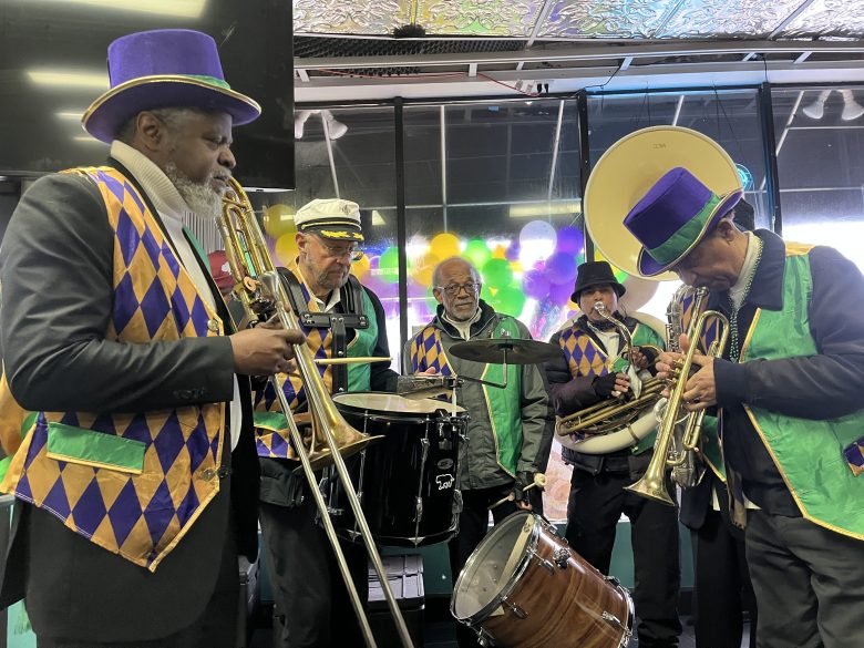 A jazz band dressed in colorful Mardi Gras-themed outfits performs indoors with trombones, a sousaphone, and a drum set.