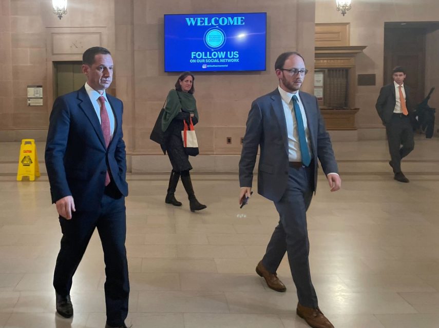 Two men in suits walk through a large indoor hallway with high ceilings; a digital welcome sign and a "wet floor" caution sign are visible in the background.