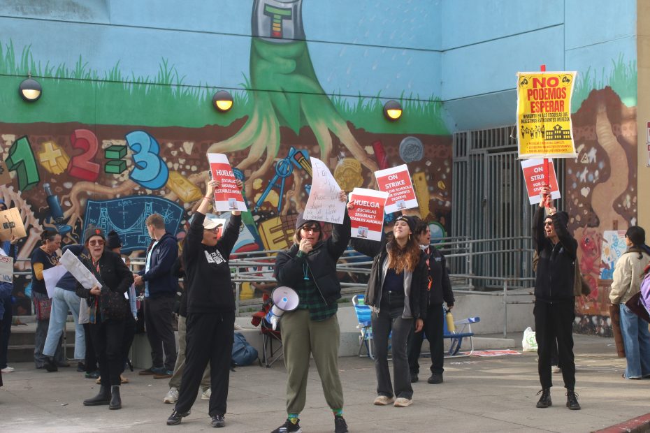 A group of people protest outside a building with a mural, holding signs with messages such as "ON STRIKE" and "NO PODEMOS ESPERAR.