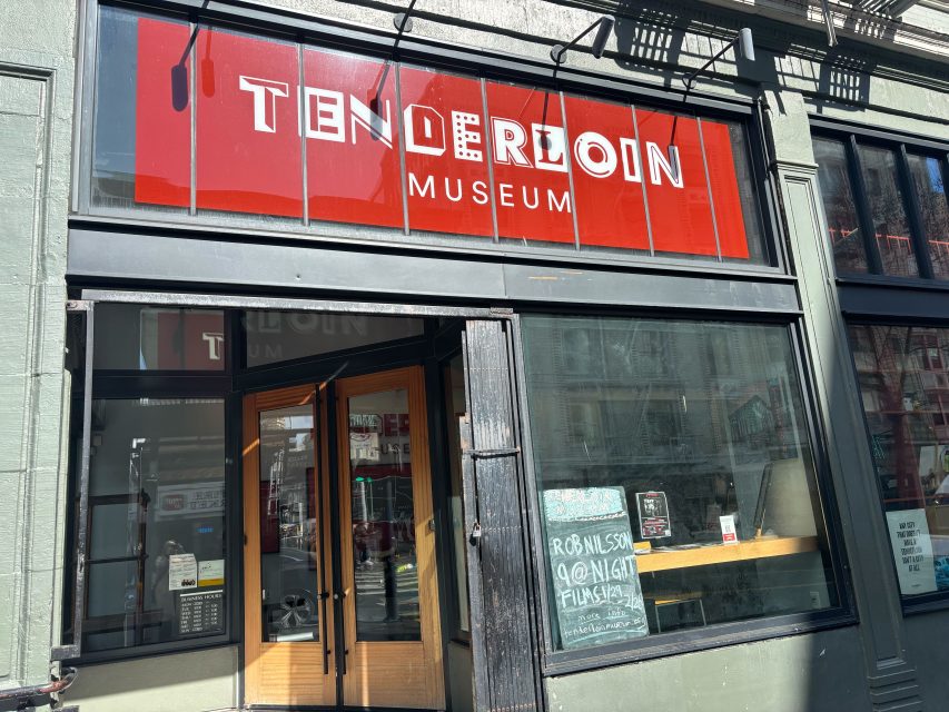 Street-level view of the entrance to the Tenderloin Museum with a bright red sign above and a sidewalk chalkboard near the door.