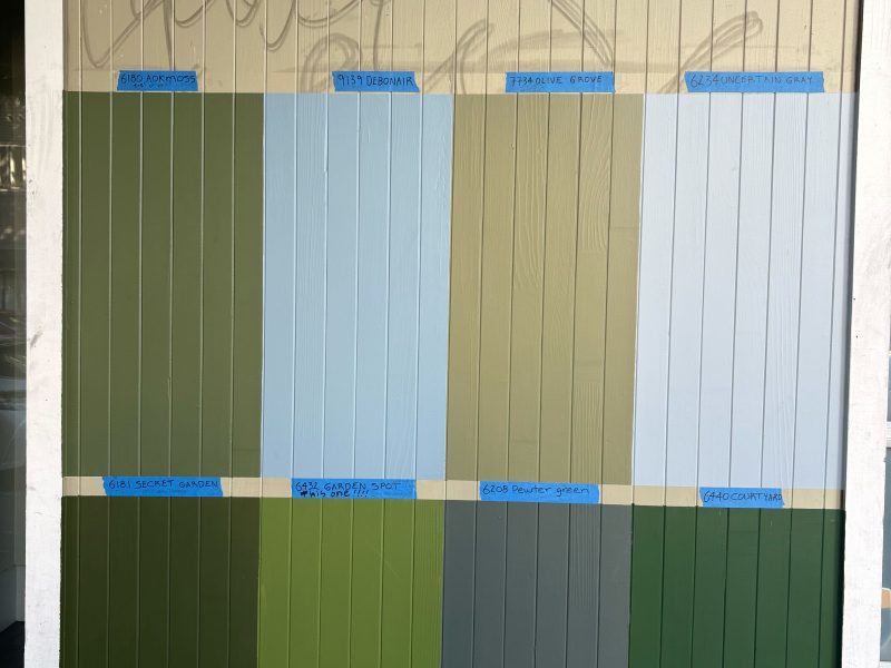A wall with vertical panels displays nine rectangular paint swatches in various shades of green and light blue, each labeled with blue painter's tape.
