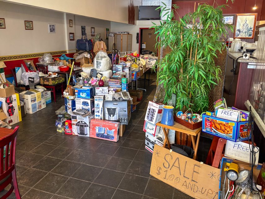 A room filled with assorted household items and appliances for sale, with boxes and a handwritten sign reading “SALE $1.00 AND UP.” A “FREE” box sits on a table near a large potted plant.