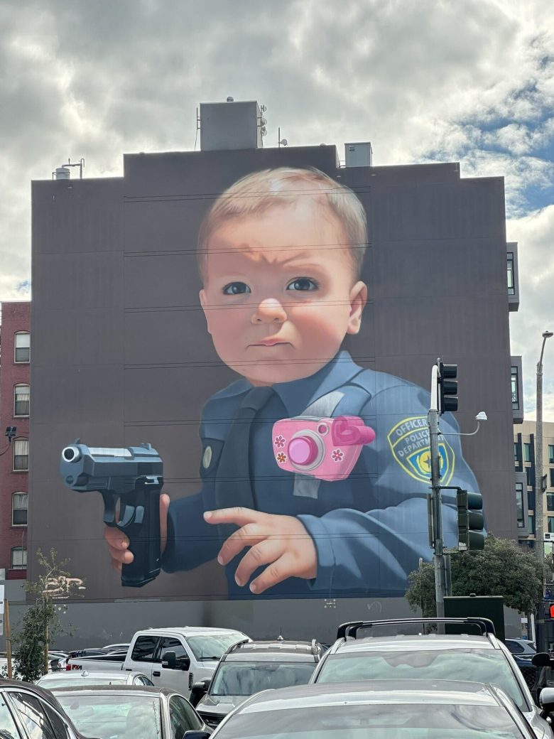 A mural on a building shows a baby in a police uniform holding a toy camera and a handgun, with parked cars and traffic lights in the foreground.