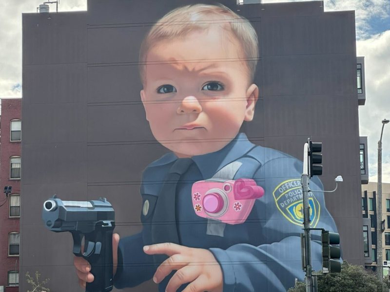 A mural on a building shows a baby in a police uniform holding a toy camera and a handgun, with parked cars and traffic lights in the foreground.