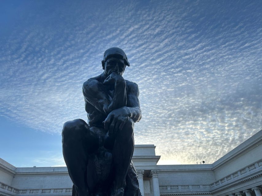 Bronze statue of a seated man with his chin resting on his hand, set against a cloudy sky and classical building facade.