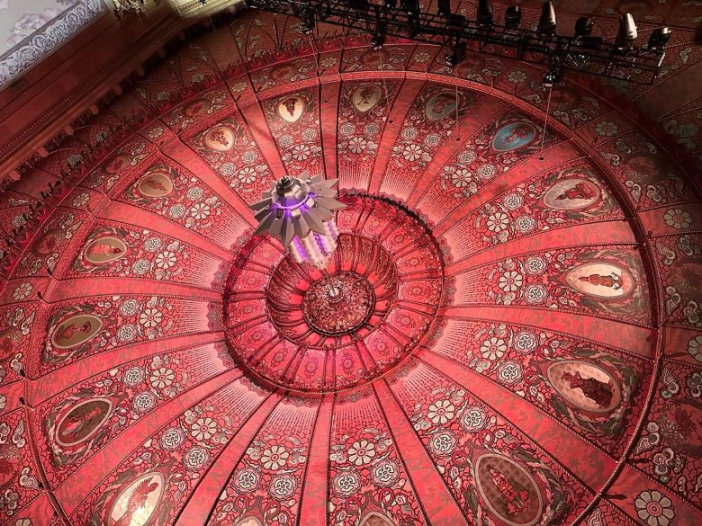 Circular, ornate red ceiling with intricate floral patterns, portrait panels, and a central chandelier, viewed from below.