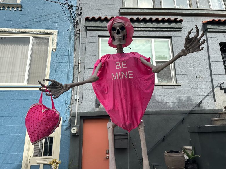 A tall skeleton figure dressed in a pink heart-shaped costume with "BE MINE" text, holding a pink polka dot purse, stands outside a gray and blue building.