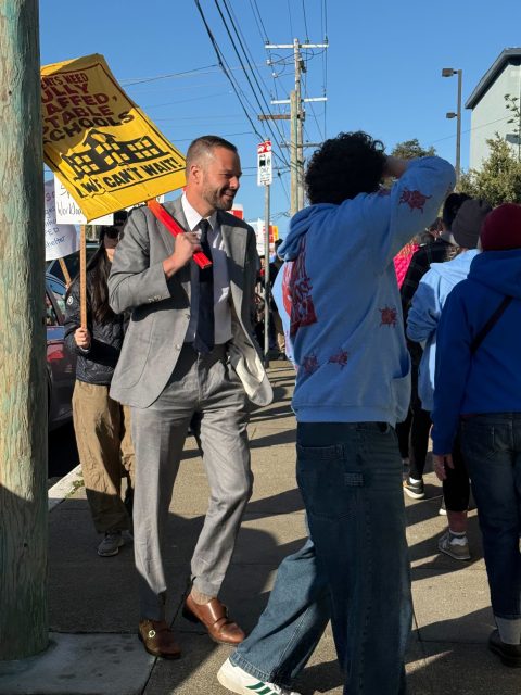 A group of people walk on a sidewalk during a protest. One person holds a yellow sign that reads, "We Want Safe Fully Staffed Schools. We Can't Wait!.