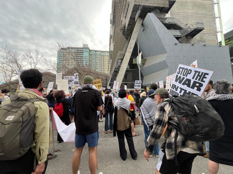 A crowd of protesters stand outside a modern building holding signs, including one that reads “STOP THE WAR.”.
