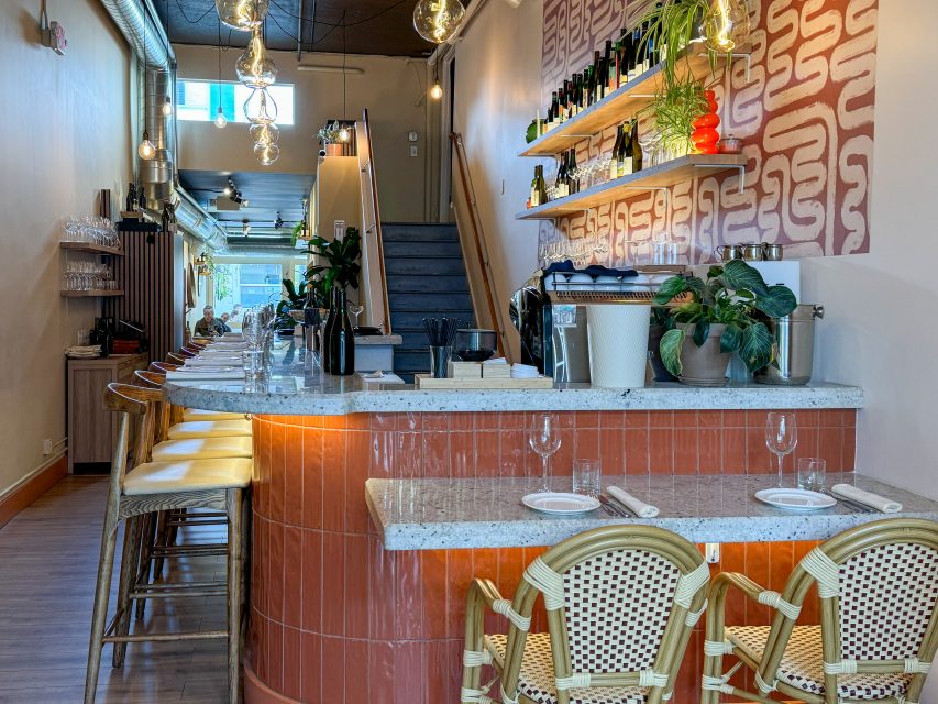 A modern restaurant interior with a curved bar, high stools, set tables, hanging lights, wall shelves with bottles, plants, and a staircase leading upstairs.