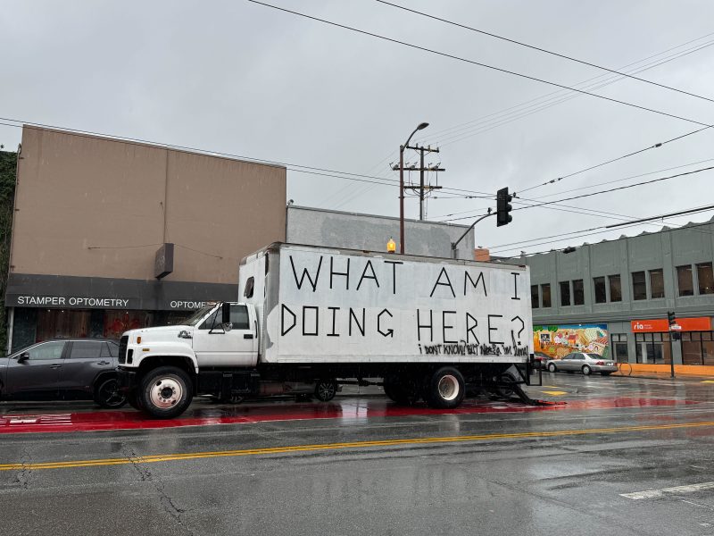 A white box truck with "WHAT AM I DOING HERE?" painted in large black letters is parked on a rainy street near shops and a mural.