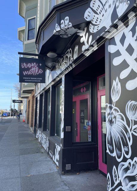 Exterior of Iron Mettle Strength Training Studio with black and white floral mural, pink doors, and a sidewalk view under a clear blue sky.