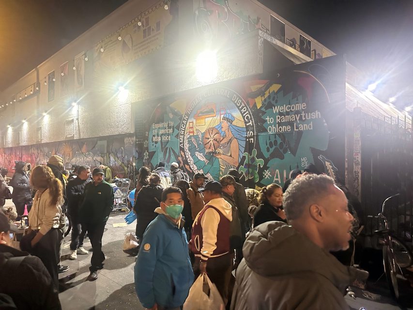 A crowd gathers at night in front of a brightly lit mural that reads "Welcome to Ramaytush Ohlone Land" in an urban setting.