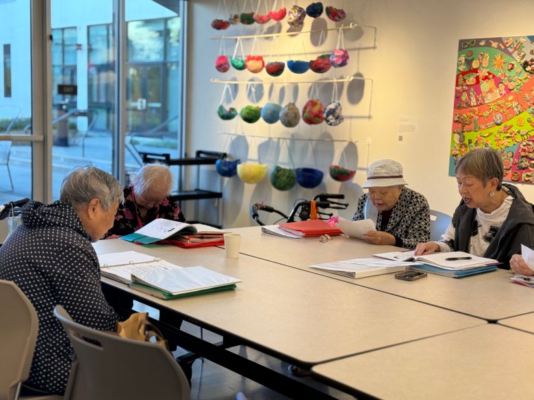 Four elderly women sit at a table in a bright room, reading and writing in notebooks, with colorful art displayed on the wall behind them.