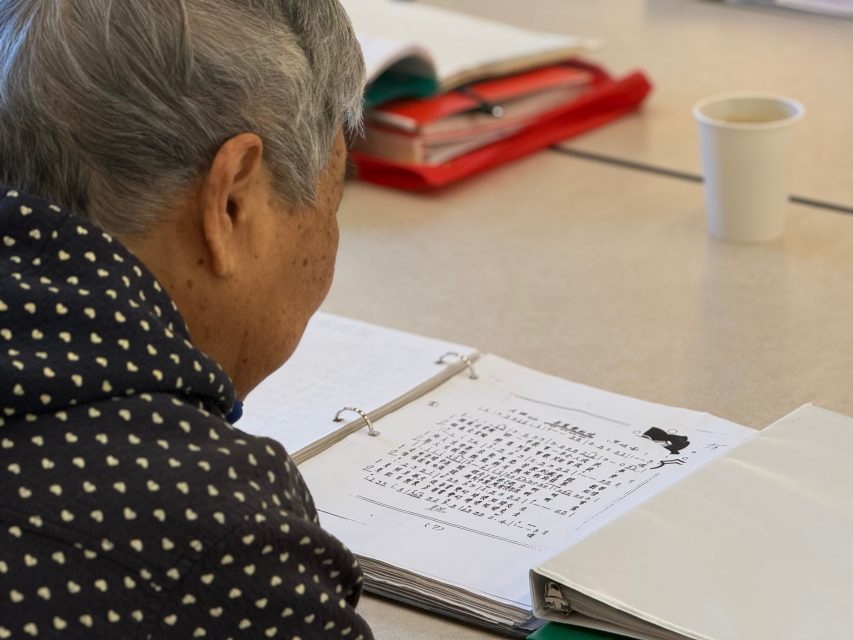 An older person with gray hair sits at a table, reading a binder with printed text in an Asian script. A paper cup and folders are on the table.