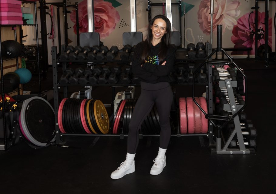 Woman standing and smiling in a gym, with arms crossed in front of a rack of dumbbells and weight plates.