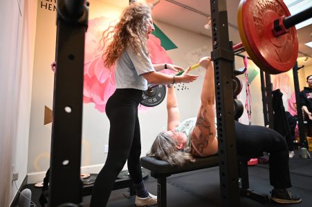 A trainer spots a person with tattoos as they lift a barbell on a bench in a gym decorated with floral wall art.