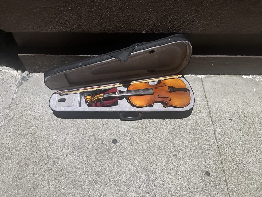A violin and bow in an open case rest on a concrete sidewalk in direct sunlight.