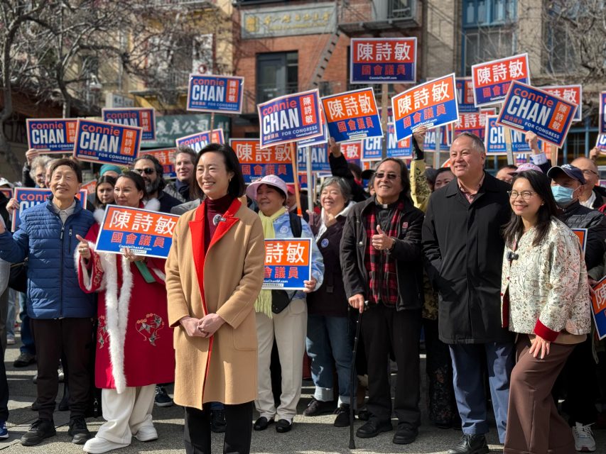 A group of people stand outdoors holding campaign signs for Connie Chan, with some wearing festive clothing and smiling at the camera.