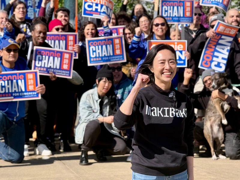 A woman stands in front of a group holding "Connie Chan for Congress" signs; she smiles and raises her fist, with supporters gathered behind her outdoors.