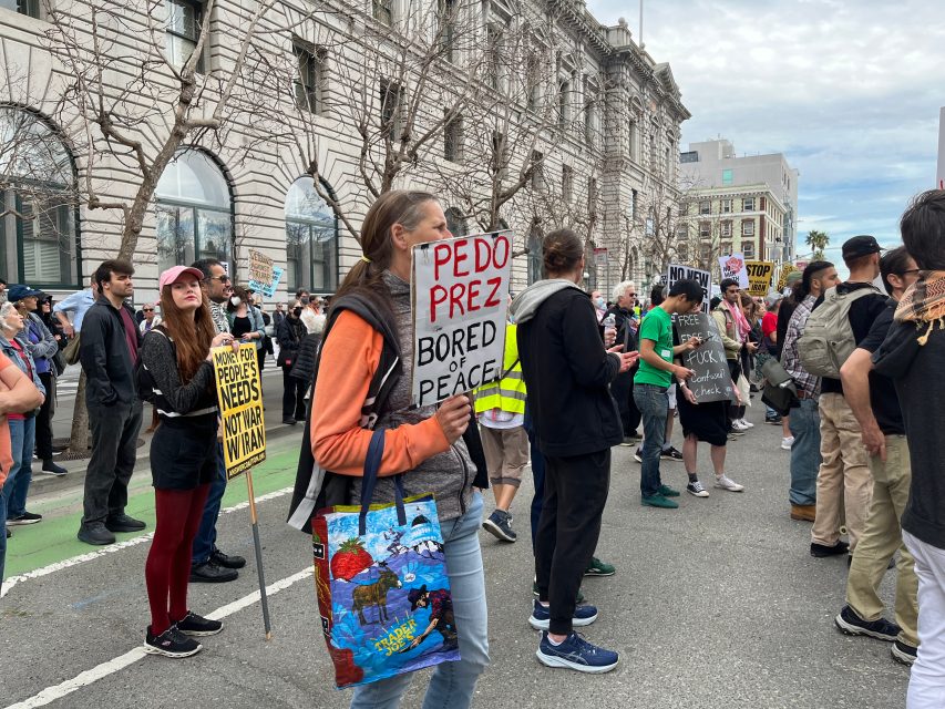 A group of people stand at a street protest holding various signs, including one reading "PEDO PREZ BORED OF PEACE" and others with slogans about people's needs and rights.
