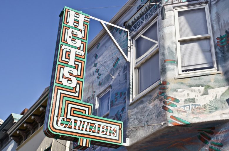 A colorful vertical sign reading "HPCSC CANNABIS" is attached to the exterior of a building with mural art and windows.