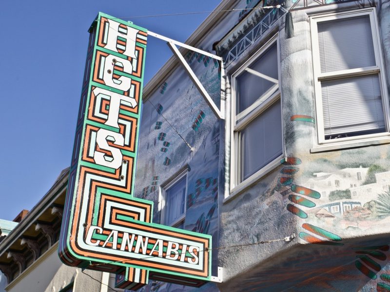 A vertical sign reading "HGS CANNABIS" hangs outside a building with a painted mural and two windows.