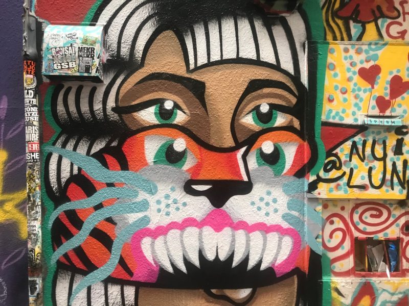 A colorful street art mural depicting a stylized face with four green eyes, white hair, and tiger-like features on a graffiti-covered wall.