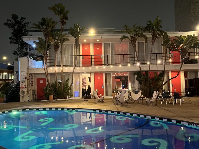 A two-story motel with red doors and balconies is lit at night, facing a swimming pool with poolside chairs and palm trees.
