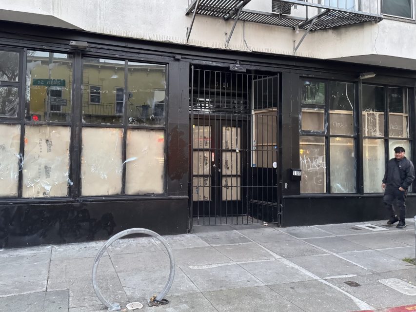 A person walks past a boarded-up storefront with barred door and graffiti on the windows, located on a city sidewalk.