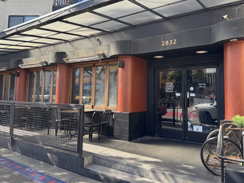 The exterior of a black and orange building with large windows, an outdoor seating area with tables and chairs, and the address number 2832 above the entrance.