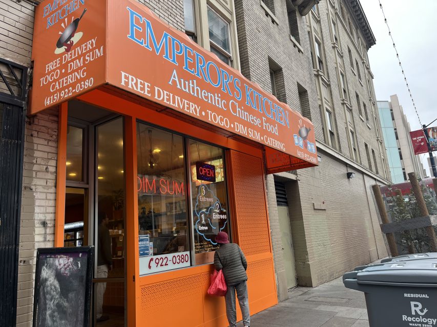 A person stands outside Emperor's Kitchen, a Chinese restaurant with a bright orange storefront offering free delivery, takeout, dim sum, and catering services.