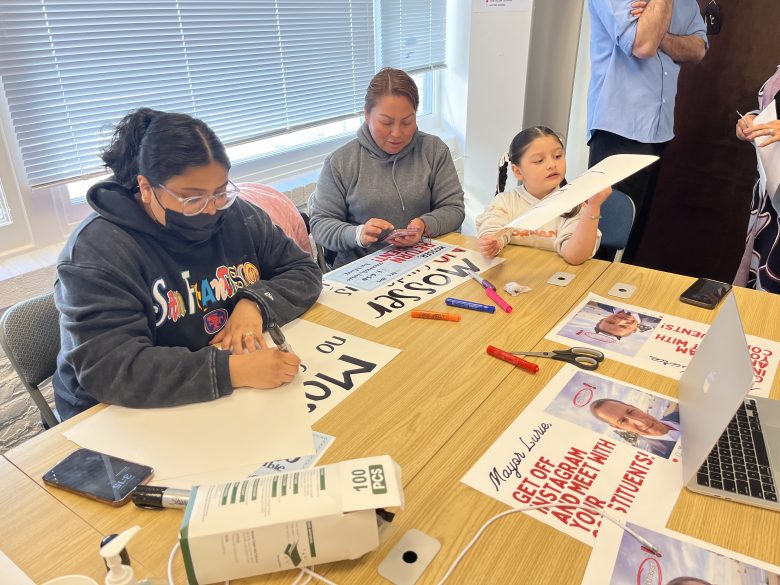Three people sit at a table making protest signs with markers and paper; printed posters and supplies are scattered on the table.
