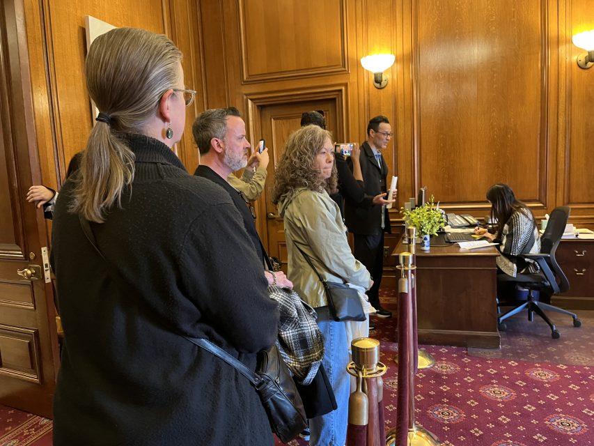 A group of people stands in an office with wood-paneled walls, watching a woman seated at a desk while another person takes photos.