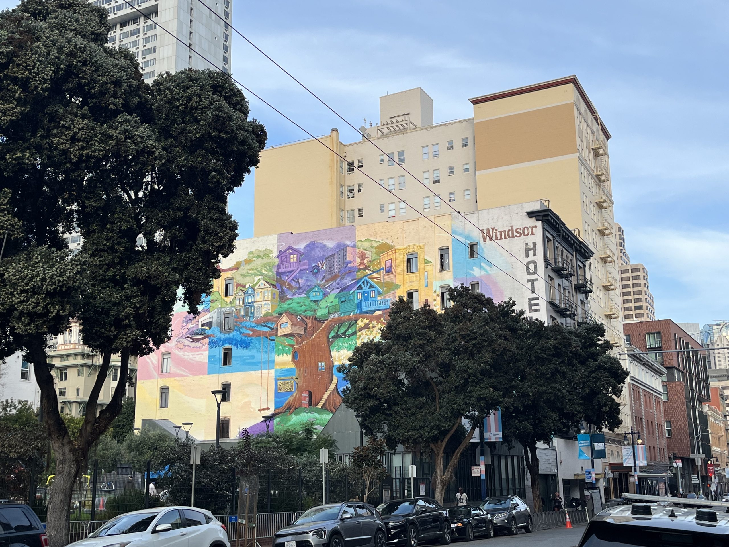 Colorful mural depicting houses and a large tree on the side of a building near Windsor Hotel in an urban area with cars parked along the street.