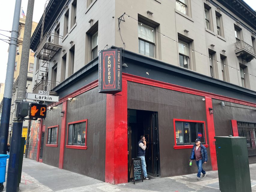 Corner view of a brown and red bar with a "Tomeroy Bar & Grill" sign; a woman stands at the entrance while two people walk past on the sidewalk at the Larkin St. intersection.