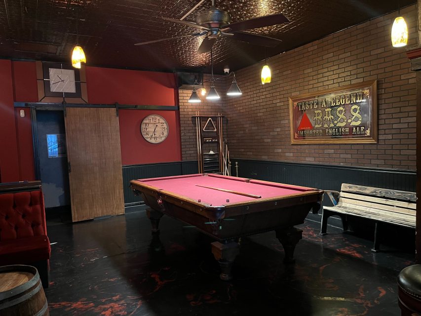 A dimly lit room with a red pool table, cues on a rack, a wooden bench, two wall clocks, and vintage signs on red and brick walls. Ceiling fan and hanging lights above.