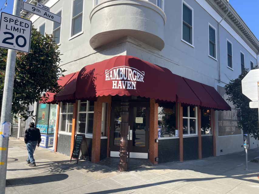 A corner restaurant with a red awning that reads "Hamburger Haven," located at a street intersection with a speed limit 20 sign and a pedestrian walking by.