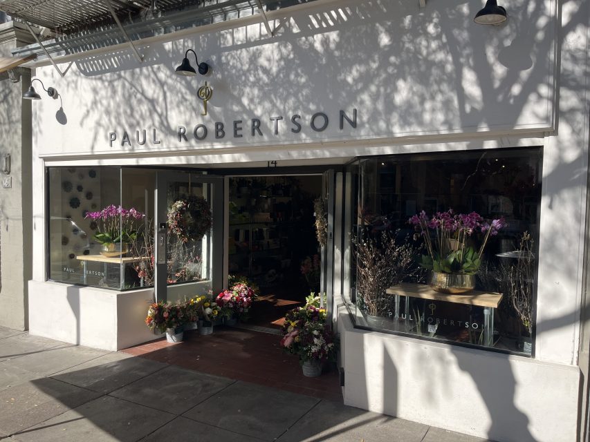 A flower shop with the name "Paul Robertson" on the facade, featuring open doors and windows with bouquets and potted plants displayed outside and inside.
