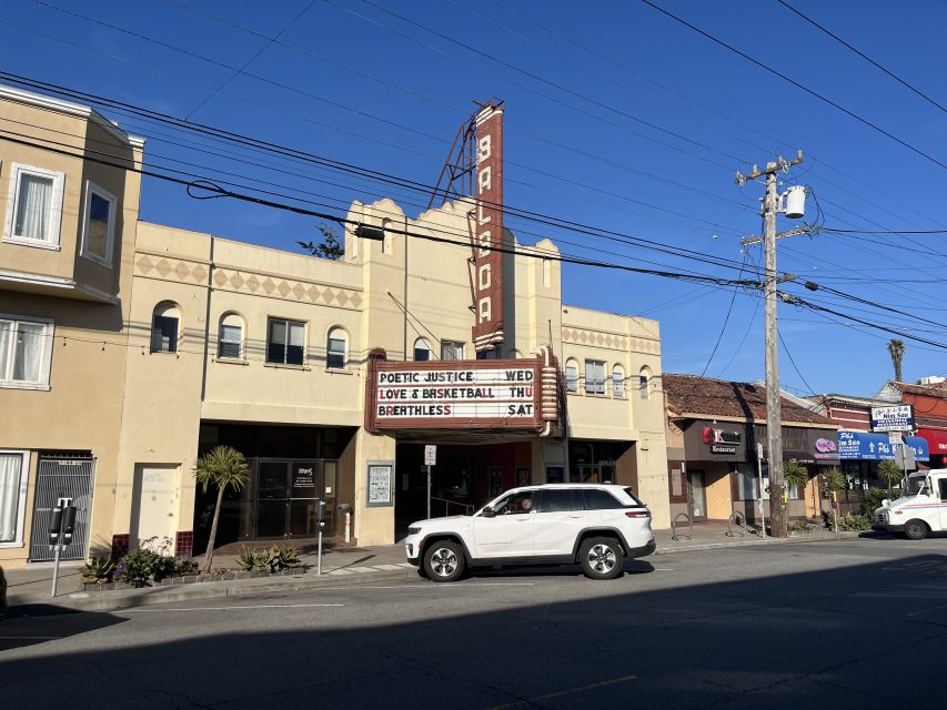 A beige Art Deco theater with a vertical โBalboaโ sign displays movie titles on its marquee; a white SUV drives by on a sunny day.